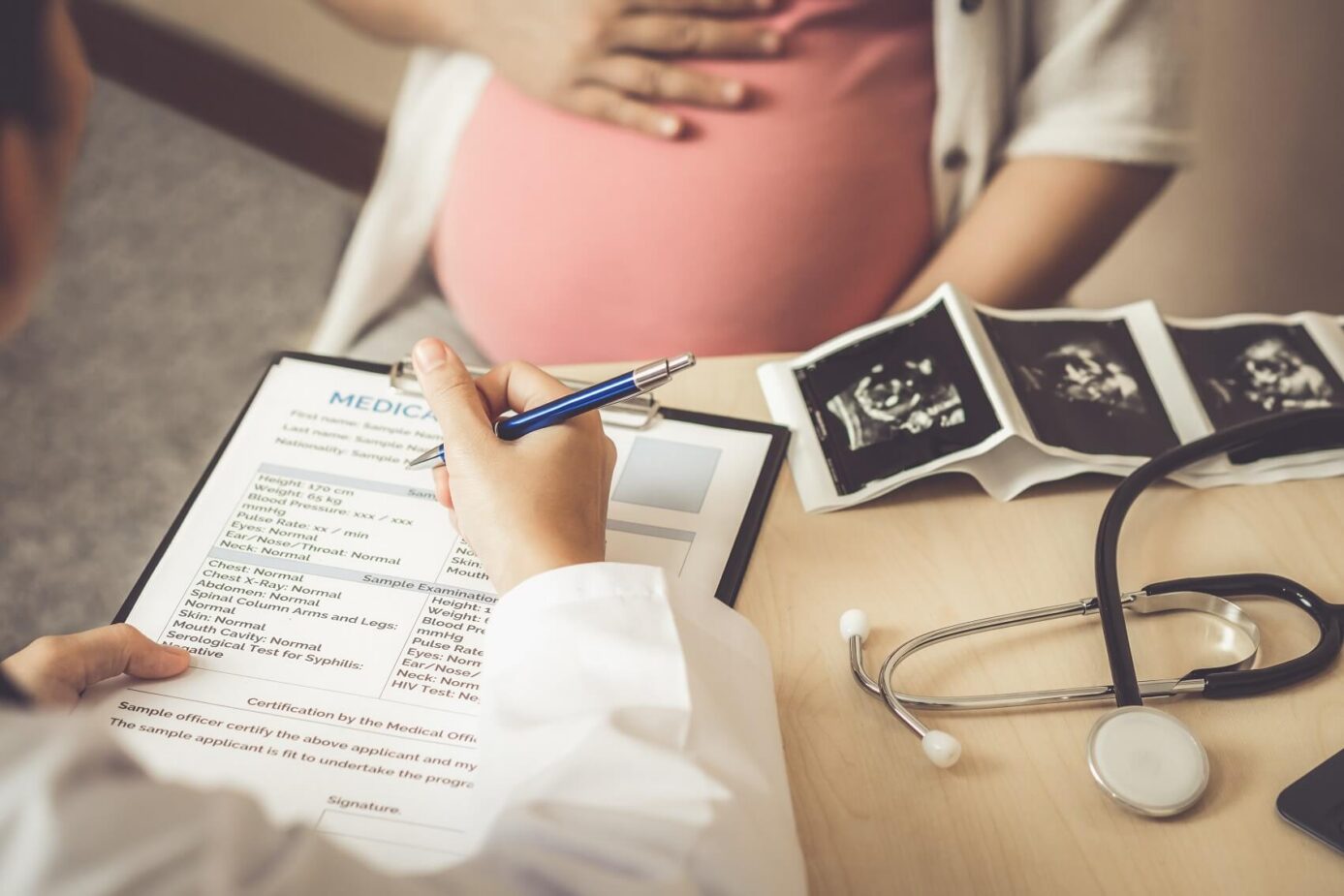 A doctor sits down at a desk while writing on a clipboard as a pregnant woman sits across the desk