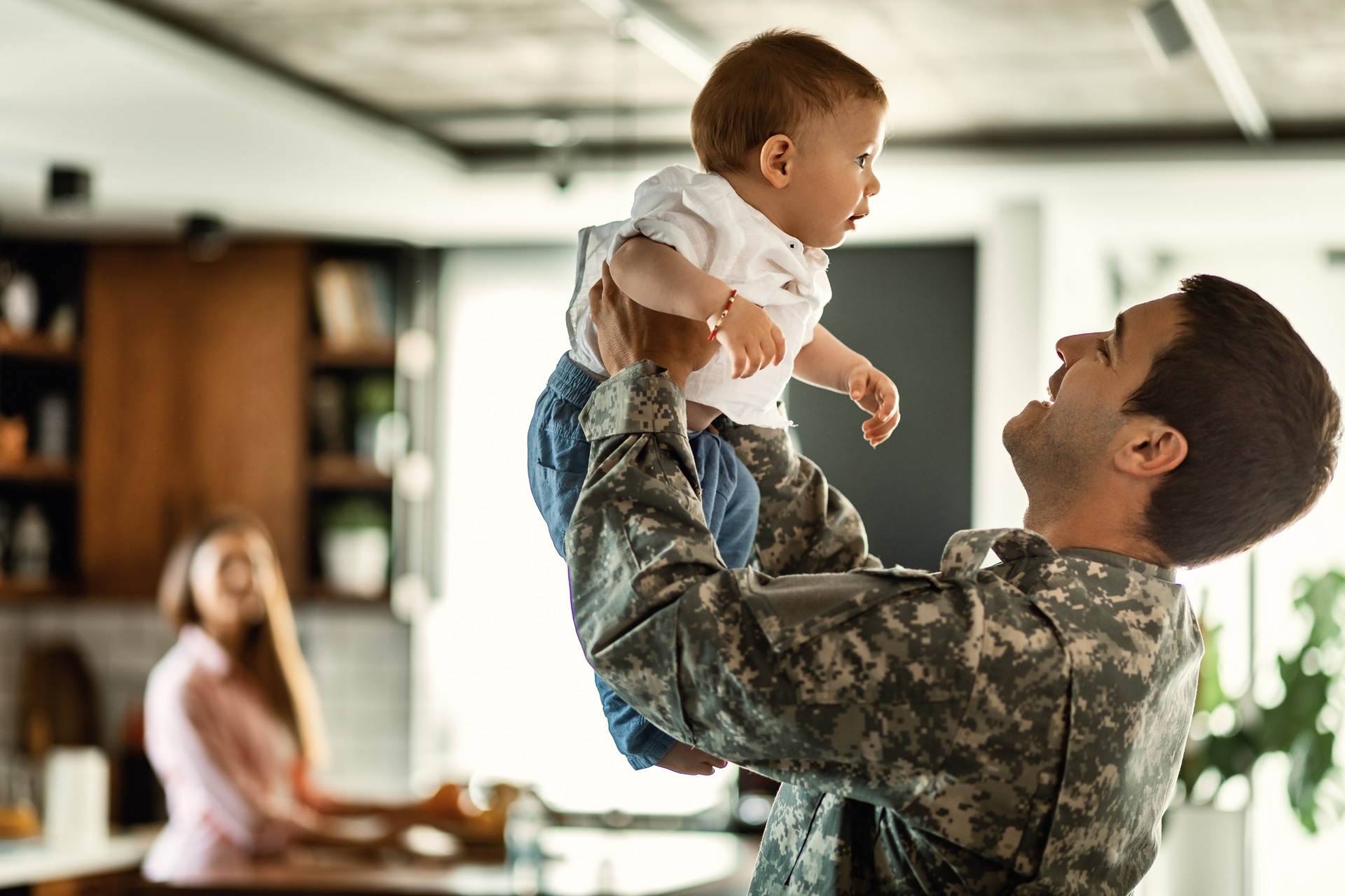 Happy army soldier playing with his baby son and having fun at home. Mother is in the background.